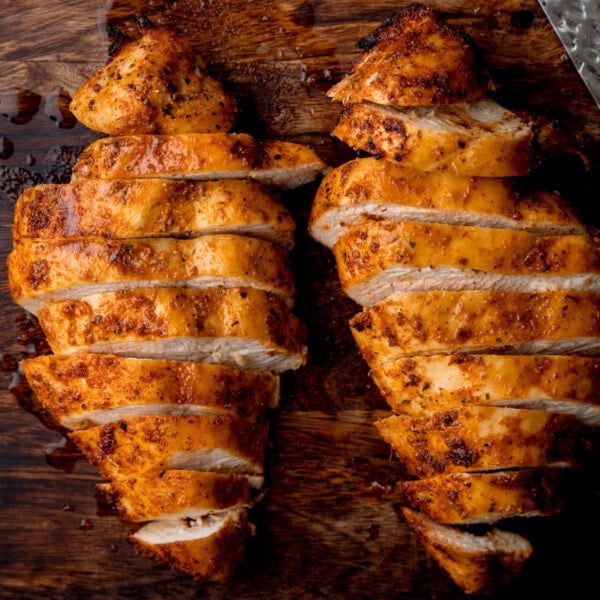 A square, overhead image of Air Fryer Chicken Breast. There are two, cooked chicken breasts next to each other in the centre of the image. The chicken breasts are cut into horizontal strips. In the top right corner of the image, you can slightly see a silver knife. This is all set on a dark, wooden background.