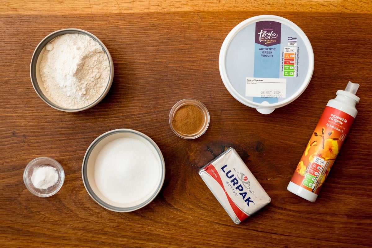 A wide, overhead shot of the ingredients for air fryer doughnut holes. The ingredients are laid out on a wooden cutting board. They are as follows: Greek yoghurt, flour, baking powder, unsalted butter, granulated sugar, ground cinnamon, and oil.