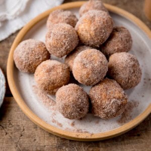 A square shot of air fryer doughnut holes in a stack on top of a white plate with a light brown rim. In the top left corner of the image, you can slightly see the corner of a white napkin. This is all set on a wooden surface.