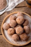 A tall overhead shot of air fryer doughnut holes in a stack on top of a white plate with a light brown rim. In the top left corner of the image, you can slightly see the corner of a white napkin and a dish of cinnamon sugar. This is all set on a wooden surface.