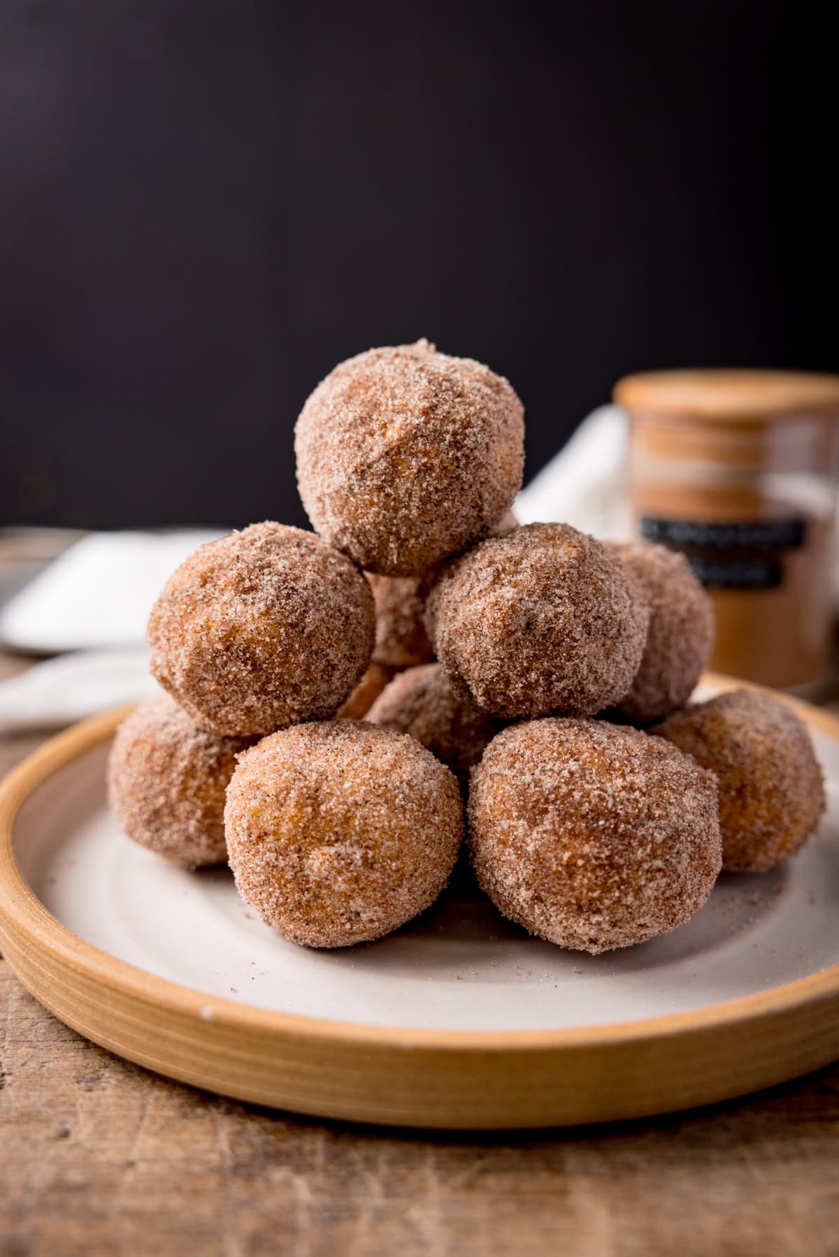 A tall shot of air-fryer doughnut holes in a stack on top of a white plate with a light brown rim. In the left of the background, you can see a clear jar, filled with ground cinnamon, with a wooden top and a black label on the side that reads cinnamon. And in the right of the background, you can see a white napkin on the surface. This is set on a wooden surface with a black background.