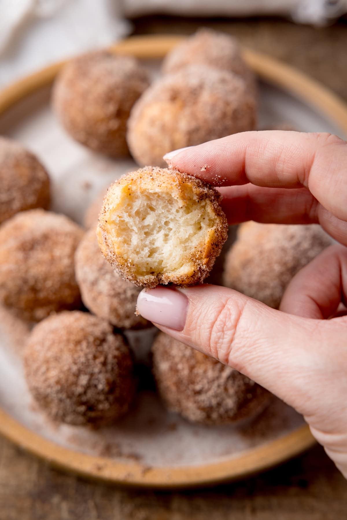 A tall shot of air fryer doughnut holes in a stack on top of a white plate with a light brown rim. In the top left corner of the image, you can slightly see the corner of a white napkin. In the centre foreground of the image, there is a single doughnut hole which has a bite taken out of it, being held by a hand that is coming from the right side of the image. This is all set on a wooden surface.