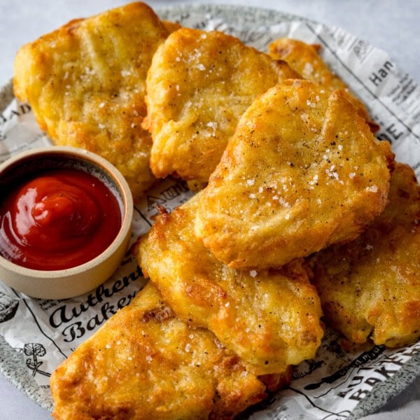 A square shot of Air Fryer Hash Browns. There are 7 hash browns in a pile on top of a plate lined with black and white newspaper parchment. There is also a small light brown pot of ketchup on the same plate, to the left of the stack of hash browns. This is all set on a plain white background.