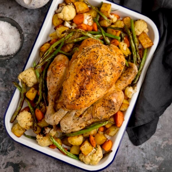 A square overhead shot of a white roasting dish filled with Air Fryer Roast Chicken Dinner, a roast chicken in the middle with potatoes, carrots and beans surrounding it. a small dark bowl filled with rock salt and a mottled blue jug filled with gravy is also next to the dish, a dark blue napkin is on the other side, set on a dark grey background.