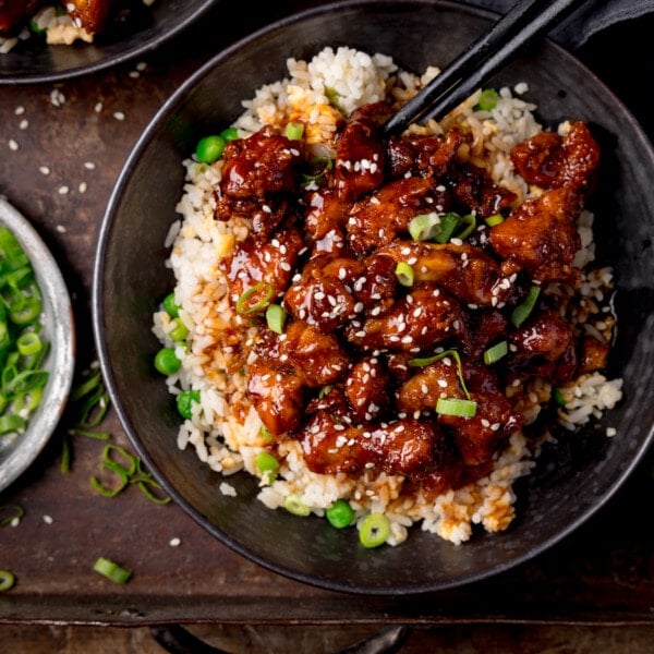 Square image of air fryer sesame chicken served on a bed of egg fried rice in a black bowl. The sesame chicken is topped with sesame seeds and spring onions. There is a pair of black chopsticks sticking out of the bowl. The bowl is on a dark background. There is a small plate with spring onions on the the left of the frame.