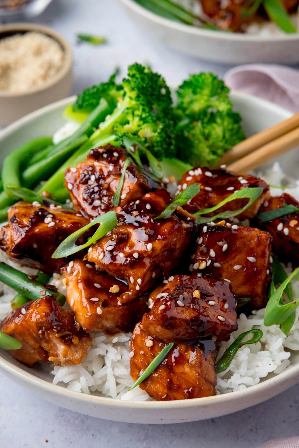 A tall photo of Air Fryer Teriyaki Salmon served on white rice, with broccoli, and topped with sesame seeds and spring onions in a white bowl. There is a pair of wooden chopsticks sticking out of the dish to the right. In the top left of the background, there is a cream dish filled with white sesame seeds. On the top right of the background, you can see a white napkin and you can slightly see another white bowl of Air Fryer Teriyaki Salmon. This is all set on a white surface.