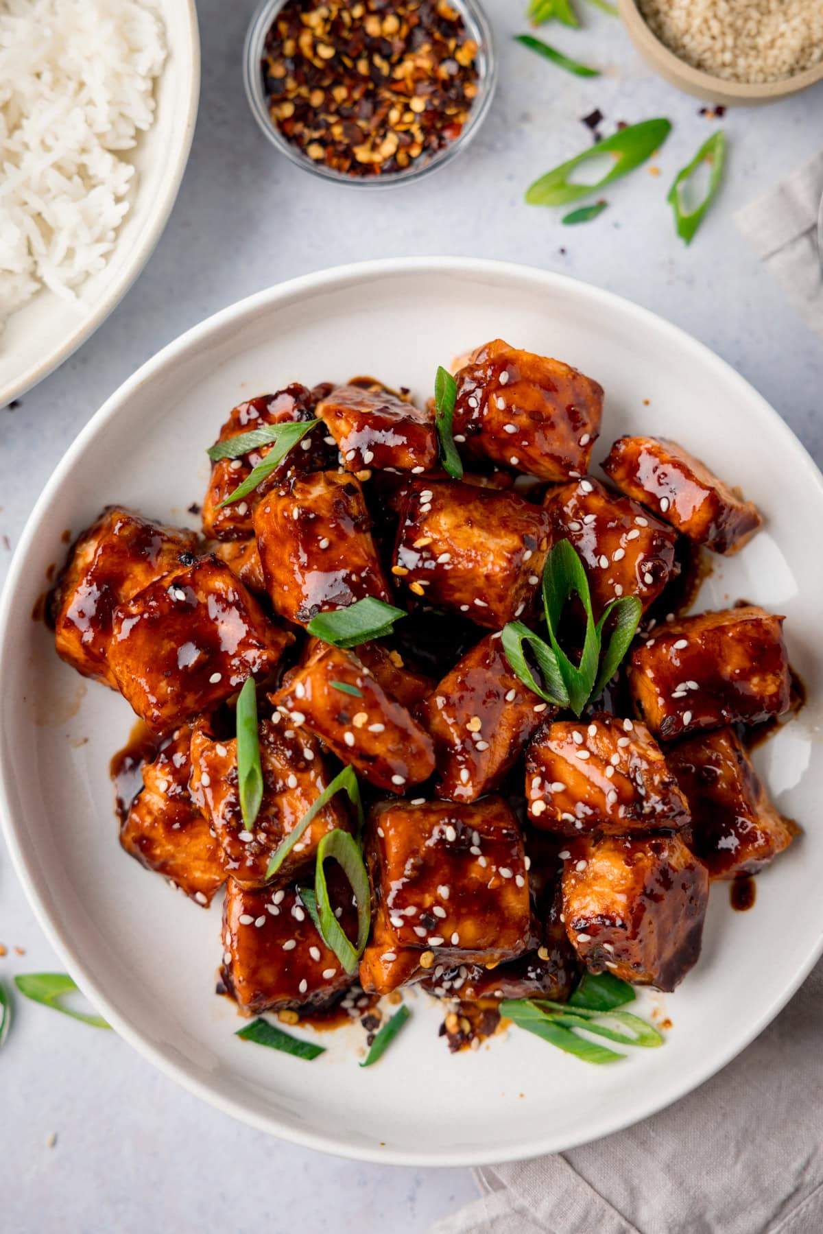 A tall overhead shot of Air Fryer Teriyaki Salmon in a white bowl, with sesame seeds and spring onions on top and scattered around the surface. On the top left of the image you can see a white bowl of cooked white rice, and next to it a small clear dish of dried chilli flakes. And on the top right, there is a cream dish of sesame seeds. There is also a napkin beneath the main bowl of Air Fryer Teriyaki Salmon. This is set on a white background.