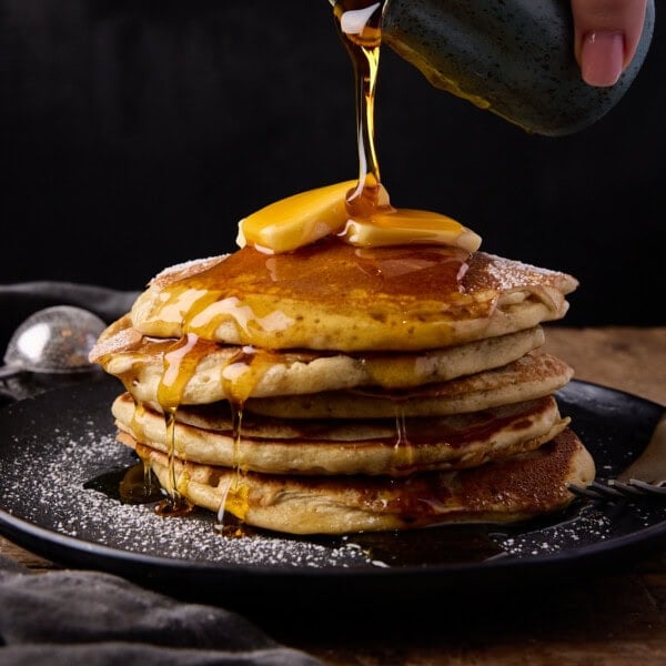 A square shot of a stack of six fluffy pancakes on top of a black plate. On top of the stack is two slices of butter and there is syrup being poured from a dark green jug above the pancakes. In the left of the background, there is a black napkin tucked beside the plate, on top of the napkin is a small circular sieve just behind the plate. This is all on a wooden surface with a black background.