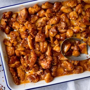 A square, overhead shot of Baked Honey Garlic Chicken. The honey garlic chicken is in a large white tray, with a dark blue rim. To the right of the image, you can slightly see a large silver spoon resting in the tray with the chicken, there are a few pieces of chicken in the spoon. You can see to the left of the background, that there are a few dried chilli flakes scattered around the tray, and to the bottom right of the image, you can see a white napkin tucked underneath the tray. This is all set on a light grey background.