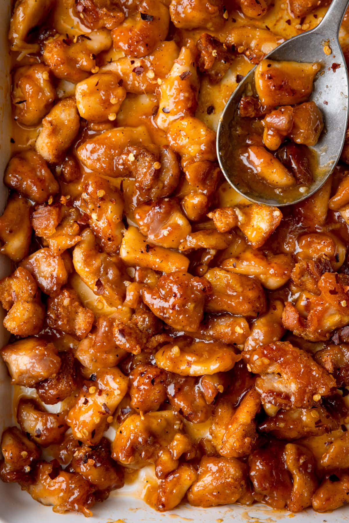 A tall, closeup shot of Baked Honey Garlic Chicken. The picture of the tray of baked chicken fills the image so the background cannot be seen. The chicken is in a white tray, and in the top right corner of the image, you can see a silver spoon resting in the pan with a few pieces of chicken in the spoon.