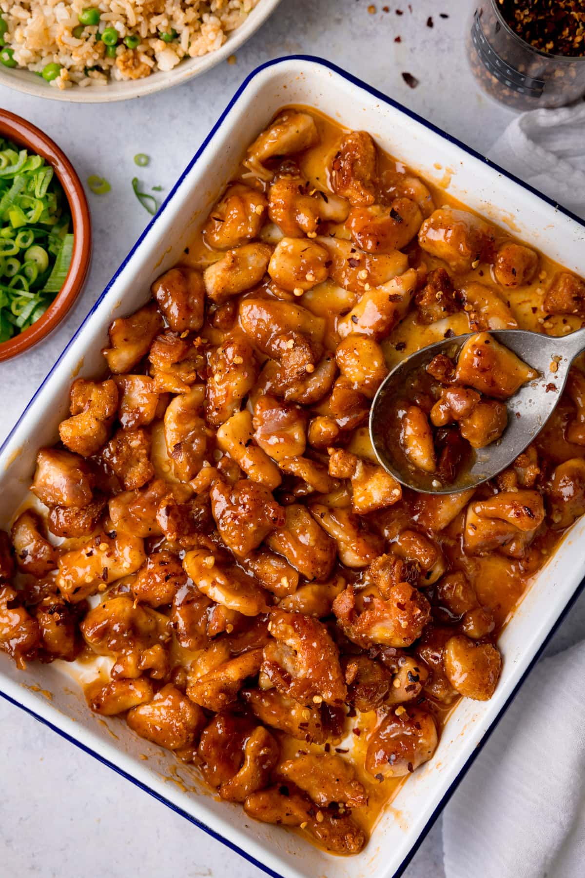 A tall, overhead shot of Baked Honey Garlic Chicken. The honey garlic chicken is in a large white tray, with a dark blue rim. The tray is in the centre of the image, at a slight diagonal. You can slightly see a large silver spoon resting in the top right corner of the tray with the chicken, there are a few pieces of chicken in the spoon. In the top left of the image, you can see the corner of a brown dish filled with whopped spring onions. Above it is a white bowl with fried rice in it, and in the top right of the image, you can see the corner of a clear container filled with dried chilli flakes. You can see to the bottom right of the image, there is a white napkin tucked underneath the tray. This is all set on a light grey background.