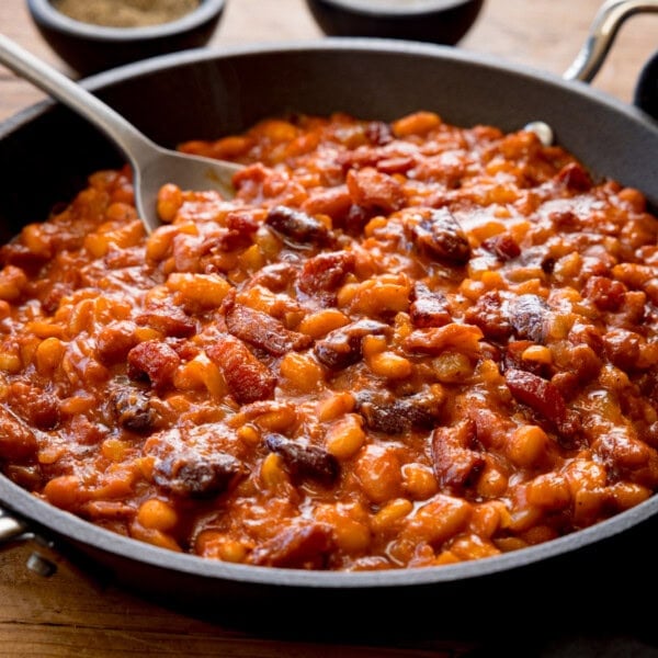 A square shot of BBQ Beans. The BBQ Beans are in a black skillet, with a silver spoon sticking out of the dish. In the top of the background, you can see two small black dishes, one with salt and one with ground pepper. This is all set on a wooden background.