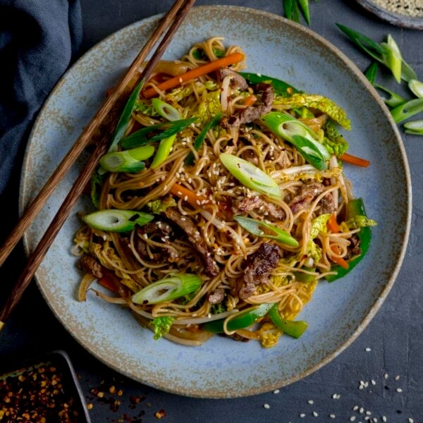 Square image of beef chow mein on a blue plate, with chopsticks resting on the plate.