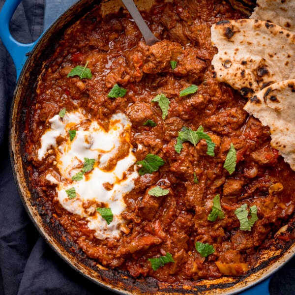 Overhead square image of Beef Rogan Josh in a blue pan. The curry is topped with chopped mint leaves, a swirl of plain yogurt and some torn chapati.