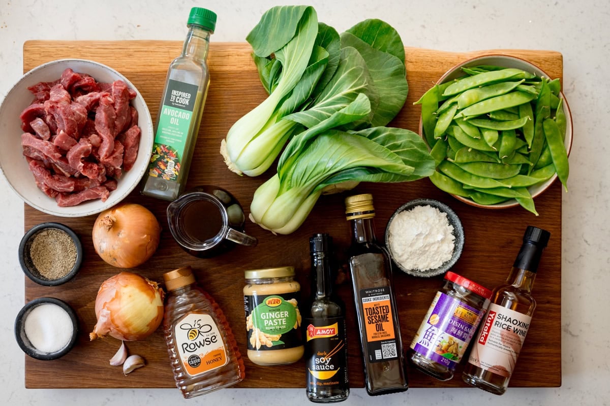 A wide, overhead shot of the ingredients for beef stir fry, they are laid out on a wooden board, which is on a white marble surface. The ingredients are as follows: steak, salt, pepper, oil, onion, mange tout, pak choi, garlic, ginger, cornflour, sesame oil, soy sauce, hoisin sauce, rice wine vinegar, honey, and beef stock.