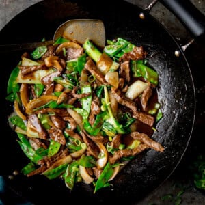 A square, overhead image of Beef Stir Fry in a black wok. The wok with the stir fry is in the centre of the image. In the right bottom corner of the image, you can see some green