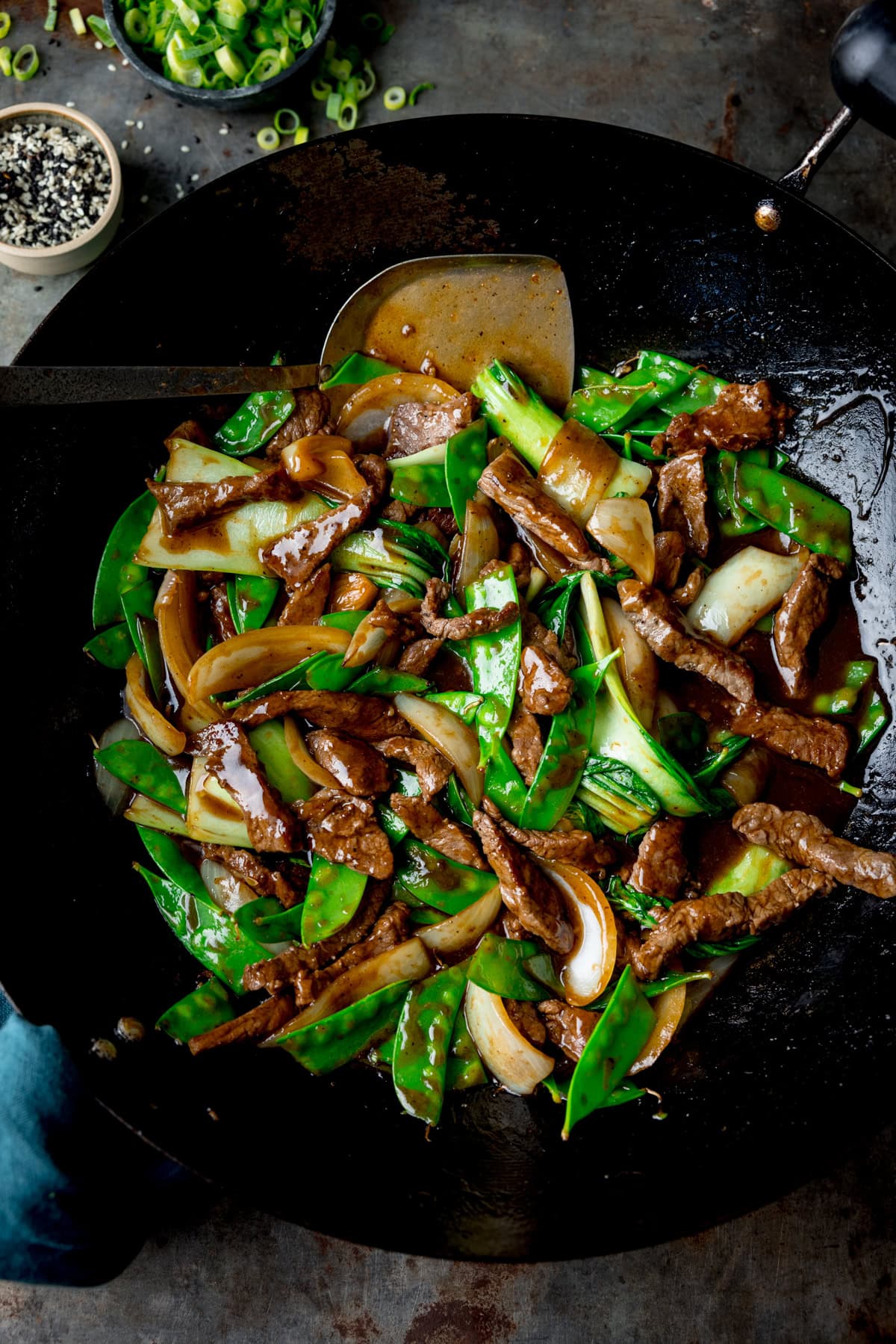 A tall, overhead image of Beef Stir Fry in a black wok. The wok with the stir fry is in the centre of the image, tucked to the left of the wok is a teal napkin. In the wok, there is a silver spatula with a wooden handle in the dish. In the top left corner of the image, there is a small black dish filled with chopped spring onions, there are also chopped spring onions scattered around it. Next to the black dish, there is a small clear dish filled with mixed sesame seeds, there are also a few sesame seeds scattered around it. This is all set on a grey background.