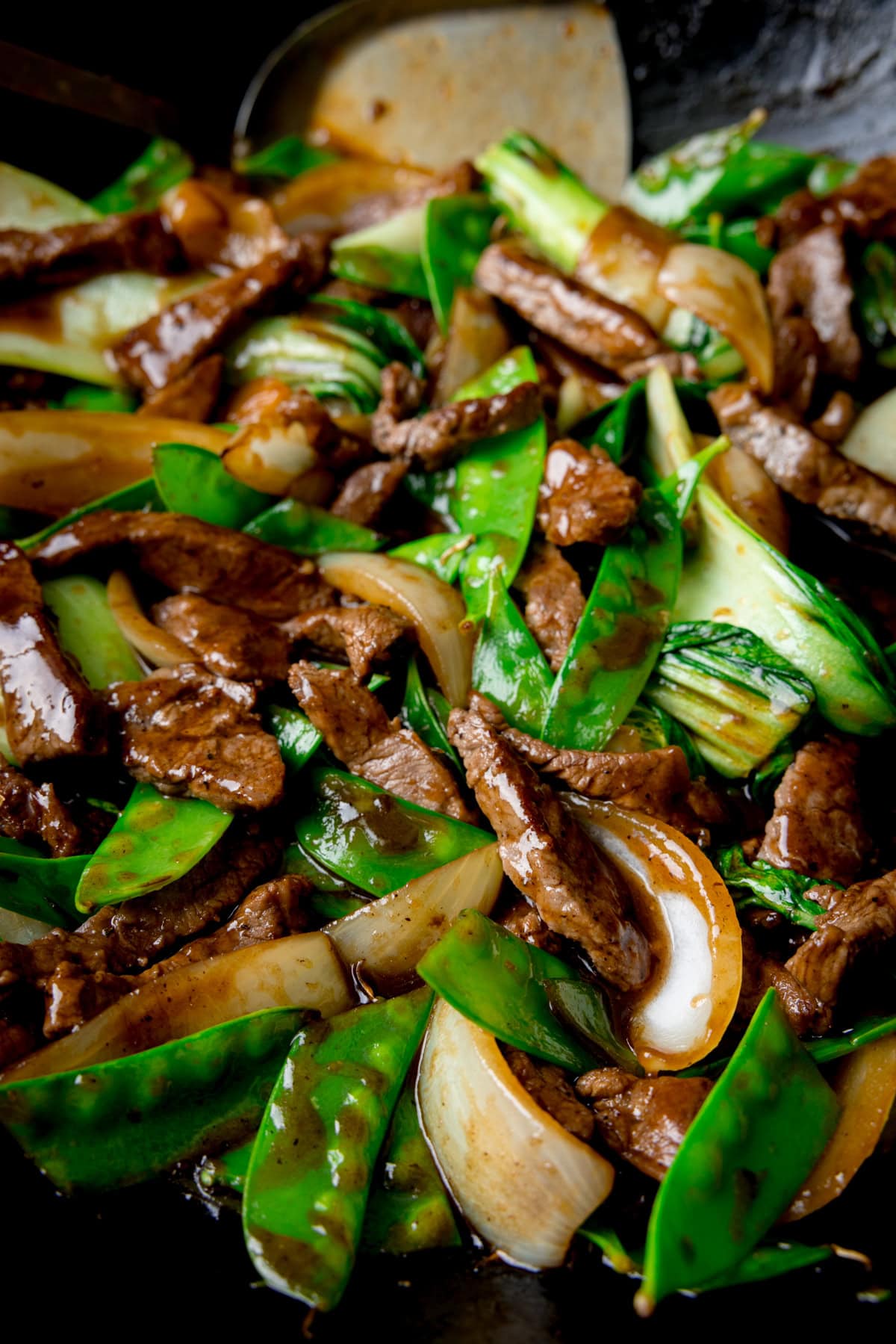 A tall, closeup shot of beef stir fry in a black wok. You can see a silver spatula sticking out of the food, resting on the side of the wok. The contents of the wok fill the screen so the background can't be seen.