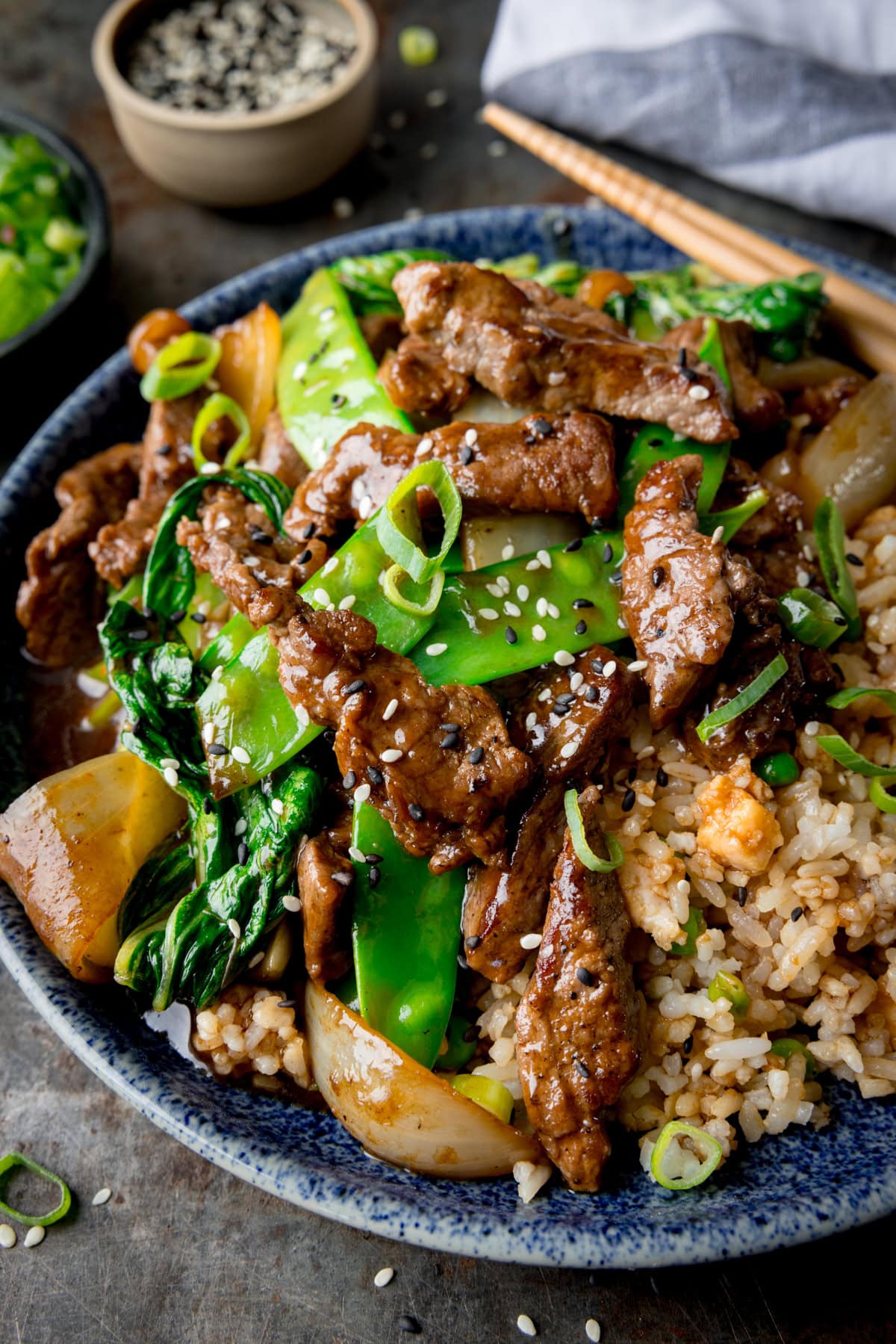 A tall image of Beef Stir Fry in a blue bowl. The bowl with the stir fry is in the centre of the image, and tucked to the top right of the bowl is a blue and white napkin. There is a pair of wooden chopsticks resting on the right side of the bowl. In the top left corner of the image, there is a small black dish filled with chopped spring onions, there are also chopped spring onions scattered around it. Next to the black dish, there is a small light brown dish filled with mixed sesame seeds, there are also a few sesame seeds scattered around it. This is all set on a grey background.