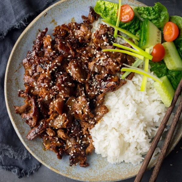 A square, overhead shot of Beef Yakiniku. The Yakiniku is on a light blue plate with light brown speckles on it. On the right side of the Yakiniku there is a small pile of white rice, and above that (still on the plate) there is a small pile of mixed chopped vegetables. On the bottom right corner of the plate, there is a pair of dark brown chopsticks resting on the plate at an angle. In the left of the background, there is a navy blue napkin tucked beneath the plate. This is all set on a light blue/grey background.