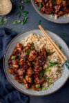 An overhead shot of a grey plate of bourbon chicken with egg fried rice, topped with spring onions and sesame seeds. The plate has a pair of wooden chopsticks lying on it. In the top right background is another grey plate of bourbon chicken, in the top right is a dish of white sesame seeds with sesame seeds and spring onions sprinkled around it. The bottom left, below the main plate of bourbon chicken, is a dark blue napkin. This is all set on a dark blue background.