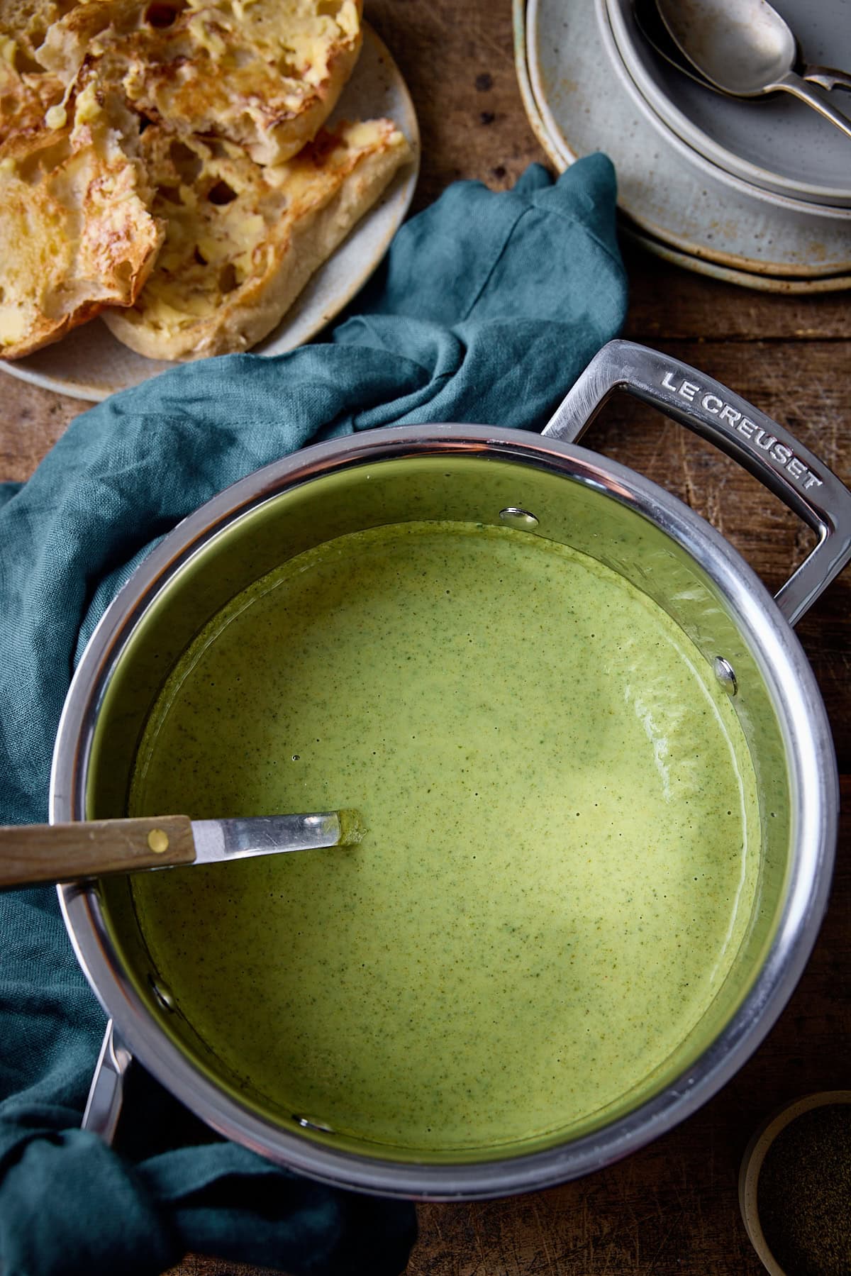 Overhead photo of broccoli cheddar soup in a stainless steel pan with a ladle sticking out. There is a green cloth and some buttered toast in the backgropund.