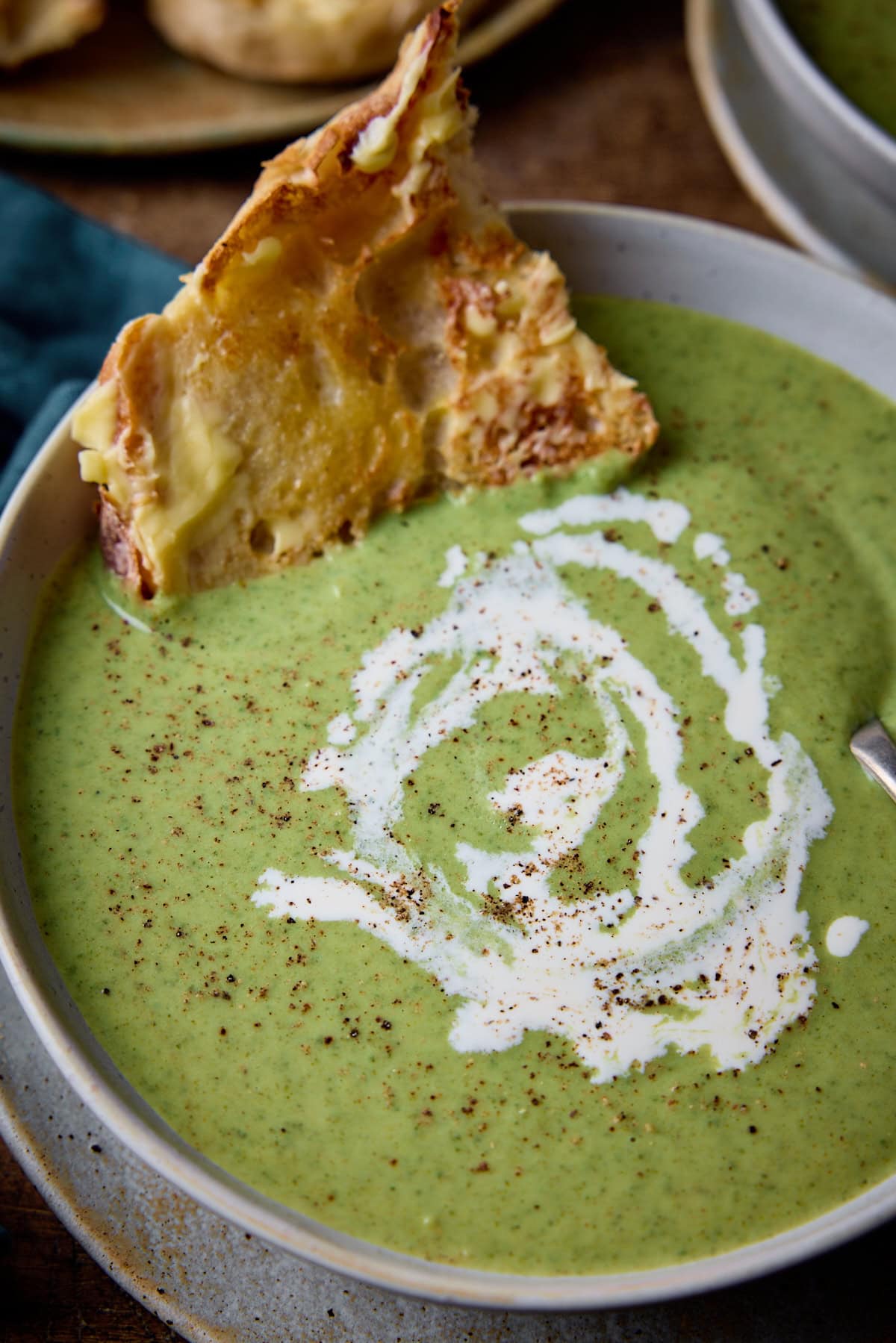 Close up picture of broccoli cheese soup in a stone light blue bowl with a piece of buttered toast sticking out of the top left corner.