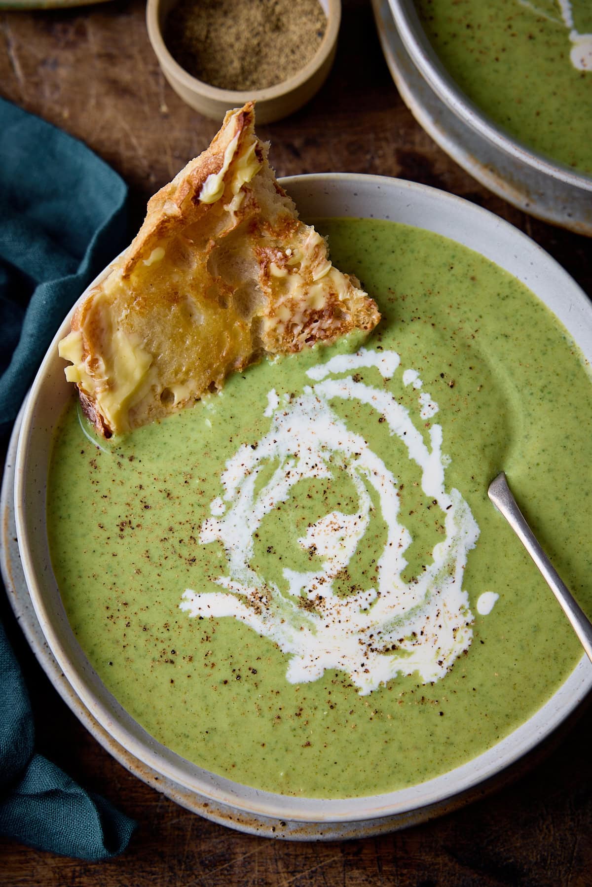 Overhead picture of broccoli cheddar soup in a stone light blue bowl with a rustic metal spoon sticking and piece of buttered toast sticking out of the soup. The soup has a swirl of cream and a little black pepper on and it's all sat on a rustic wooden table.