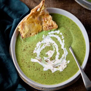 Overhead picture of broccoli cheese soup in a stone light blue bowl with a rustic metal spoon and a piece of buttered toast sticking out of the soup. The soup has a swirl of cream and a little black pepper on and it's all sat on a rustic wooden table with a green napkin in the background.