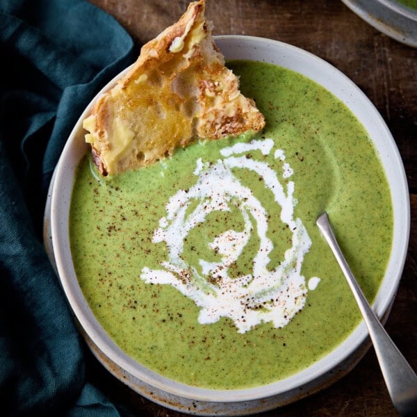 Overhead picture of broccoli cheese soup in a stone light blue bowl with a rustic metal spoon and a piece of buttered toast sticking out of the soup. The soup has a swirl of cream and a little black pepper on and it's all sat on a rustic wooden table with a green napkin in the background.