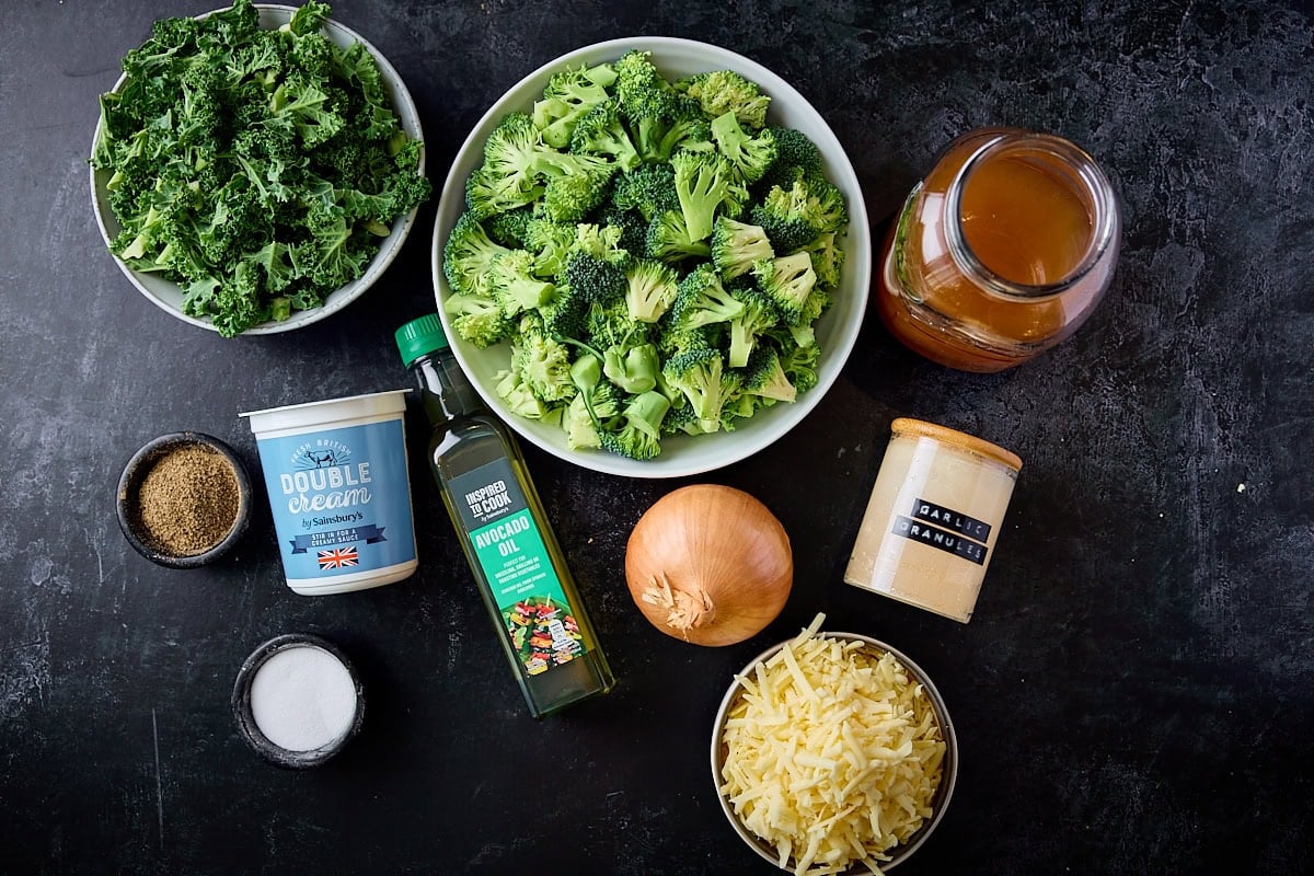 Overhead photo of the ingredients for broccoli cheese soup on a dark blue board. Key ingredients are; broccoli, kale, onion, stock, cheddar, cream and seasonings.