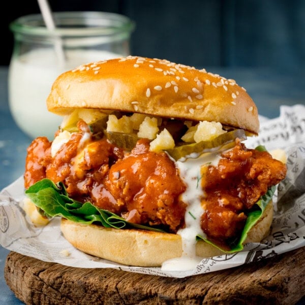 A square image of a Buffalo Chicken Burger is placed on top of a square of black and white newspaper parchment, which is on top of a dark wooden cutting board. In the left of the background, you can see a clear glass jar of homemade ranch, with a silver spoon sticking out of it. This is all set on a navy blue background.