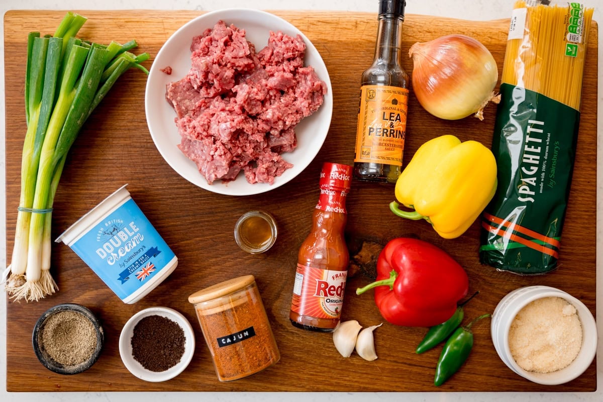 A wide, overhead shot of the ingredients for Buffalo Minced Beef Spaghetti. The ingredients are laid out on a wooden cutting board, which is on top of a white marble surface. The ingredients are as follows: spaghetti, minced beef, cajun spice mix, black pepper, beef stock cube, garlic, onion, red pepper, yellow pepper, jalapenos, spring onions, hot pepper sauce, Worcestershire sauce, cider vinegar, double cream and parmesan.