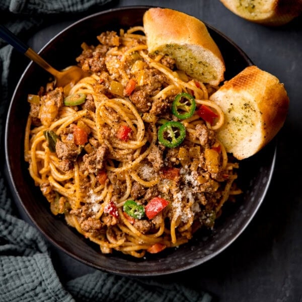 A square, overhead shot of Buffalo Minced Beef Spaghetti in a black bowl. There is a gold fork sticking out of the dish and two slices of garlic bread also in the bowl. On the left side of the image, you can see a dark grey napkin tucked around the main bowl. In the top left corner of the image, you can slightly see the corner of another piece of garlic bread on the surface. This is all set on a black surface.