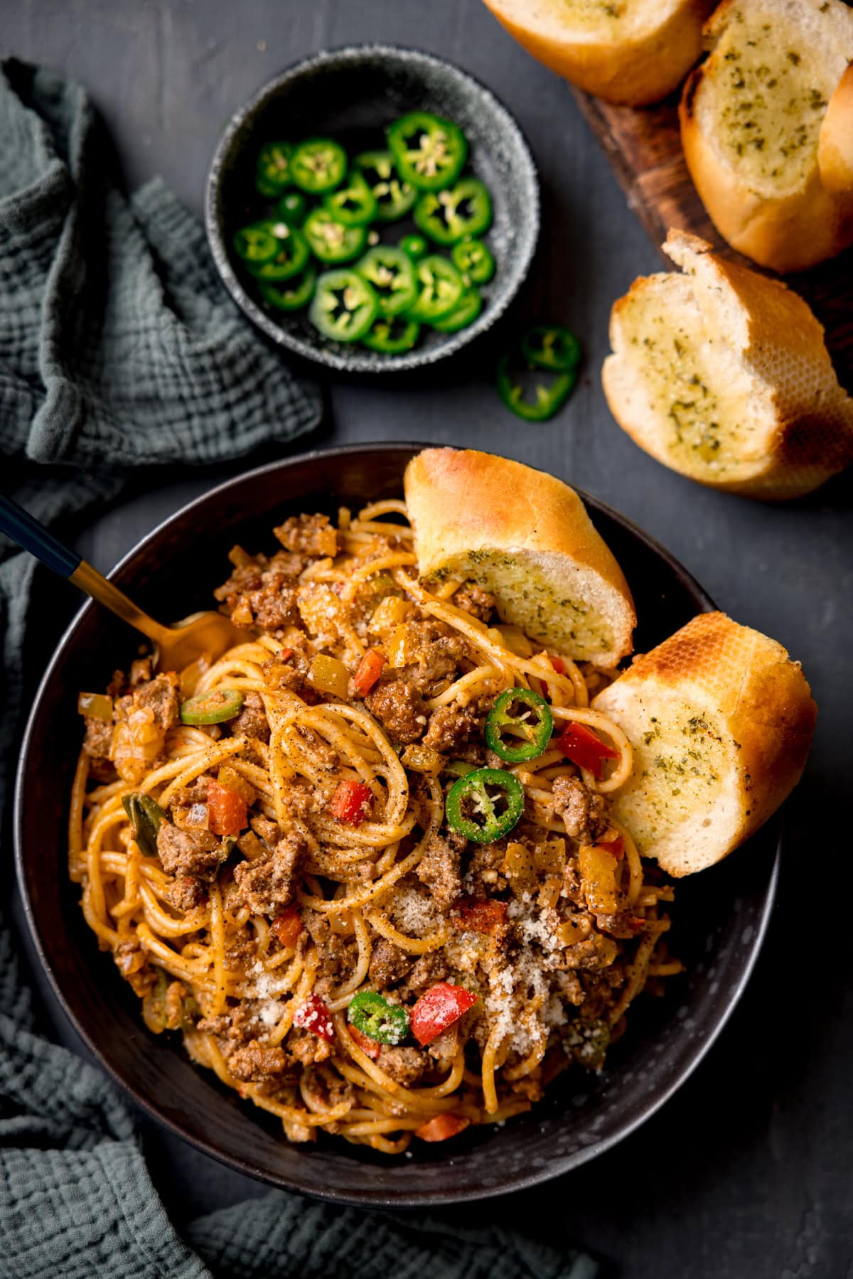 A tall, overhead shot of Buffalo Minced Beef Spaghetti in a black bowl. There is a gold fork sticking out of the dish and two slices of garlic bread also in the bowl. On the left side of the image, you can see a dark grey napkin tucked around the main bowl. In the top left corner of the image, there is a small black dish filled with sliced jalapenos, with a few sliced jalapenos scattered around it, next to it, you can slightly see the corner of three pieces of garlic bread on a wooden cutting board. This is all set on a black surface.