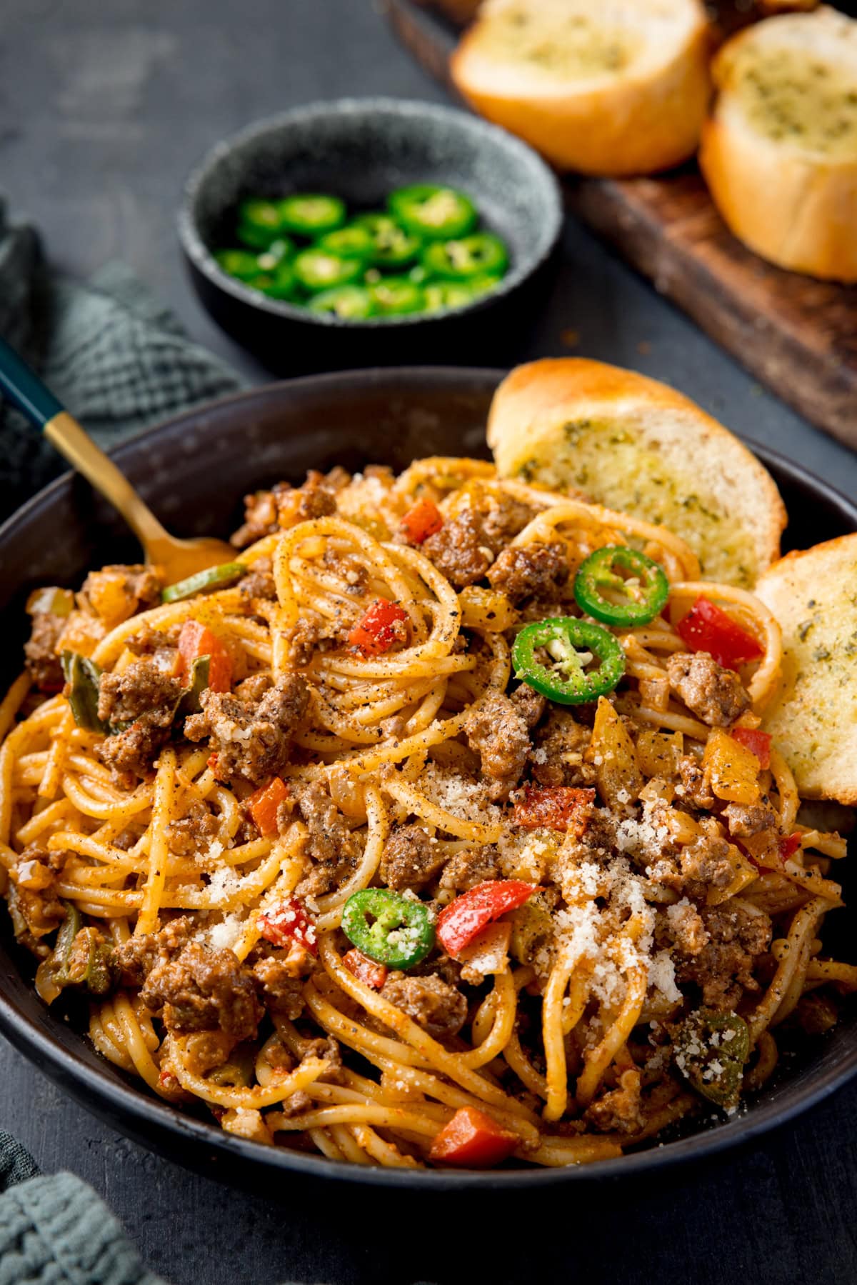 A tall shot of Buffalo Minced Beef Spaghetti in a black bowl. There is a gold fork sticking out of the dish and two slices of garlic bread also in the bowl. On the left side of the image, you can see a dark grey napkin tucked around the main bowl. In the top left corner of the image, there is a small black dish filled with sliced jalapenos, with a few sliced jalapenos scattered around it, next to it, you can slightly see the corner of two pieces of garlic bread on a wooden cutting board. This is all set on a black surface.