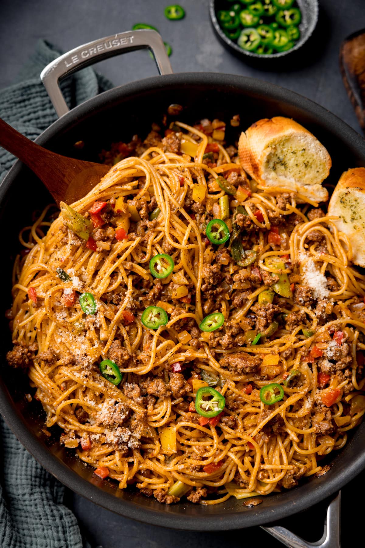 A tall, overhead shot of Buffalo Minced Beef Spaghetti in a black pan. There is a wooden serving fork sticking out of the dish and two slices of garlic bread also in the pan. On the left side of the image, you can see a dark grey napkin tucked around the pan. In the top left corner of the image, you can see a small black dish filled with sliced jalapenos, with a few sliced jalapenos scattered around it, next to it, you can slightly see the corner of a wooden cutting board. This is all set on a black surface.