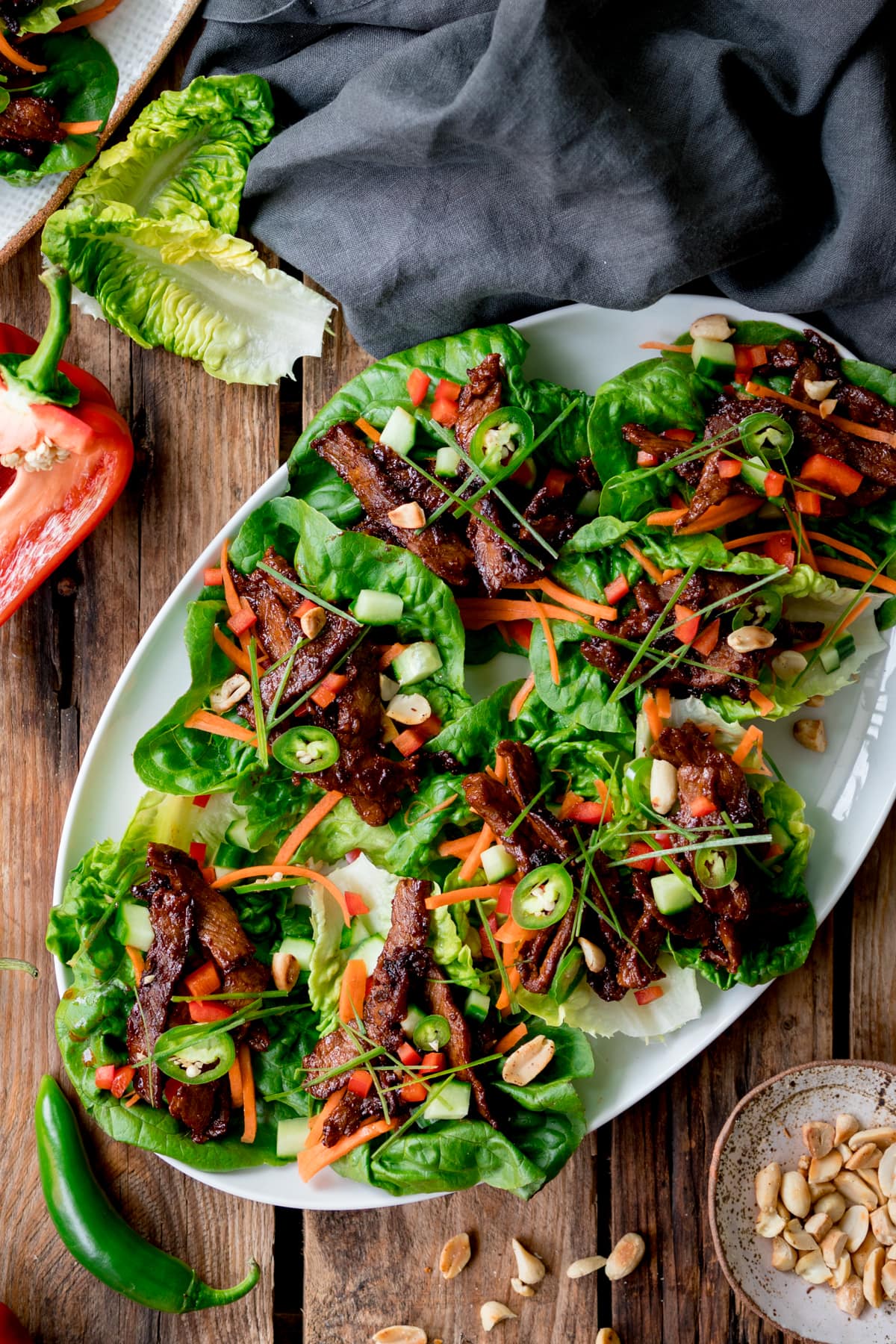 Overhead picture of a white plate full of Korean lettuce wraps all sat on a rustic wooden board with some peanuts and chillis in the background.