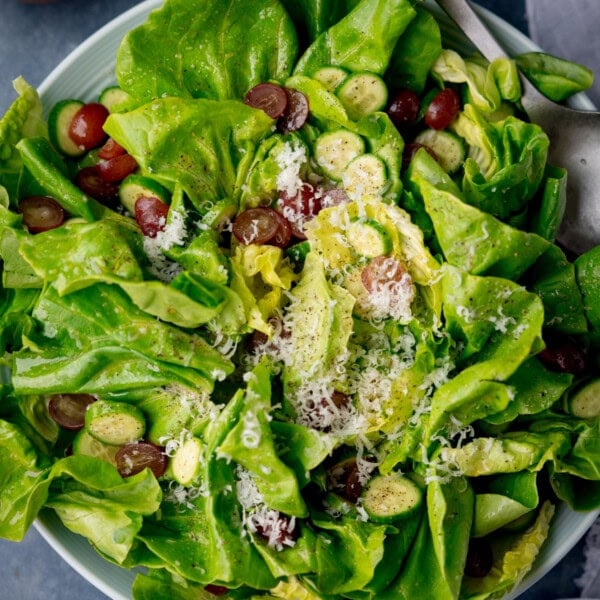 A square, overhead shot of Butter Lettuce Salad in a large white bowl. The bowl with the salad is placed in the centre of the image, in the top right corner of the bowl there are two silver metal salad salad servers sticking out of the dish. Just tucked below the right-hand side of the bowl, you can slightly see a white napkin. This is all set on a light blue/grey background.