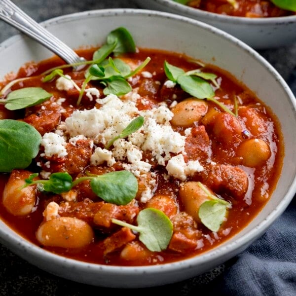 Close up square image of butterbean and chorizo soup topped with feta and watercress in a light bowl. There is a spoon sticking out of the bowl.