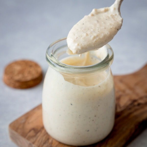 A square shot of Caesar Dressing. In the centre of the image, there is a glass jar filled with the dressing. Directly above the jar, there is a gold spoon with some dressing in it. The jar is on a small wooden board, and you can see to the left of the background there is a brown lid for the jar. This is all set on a light grey surface.