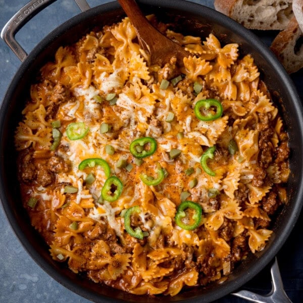 A square, overhead shot of cheeseburger pasta in a black dish with a silver handle. The pasta is topped with sliced jalapeños. In the top right corner of the dish, you can slightly see a wooden spoon sticking out of the dish at an angle. In the top right corner of the image, you can see three slices of bread stacked into a pile. This is all set in a light blue/grey background.