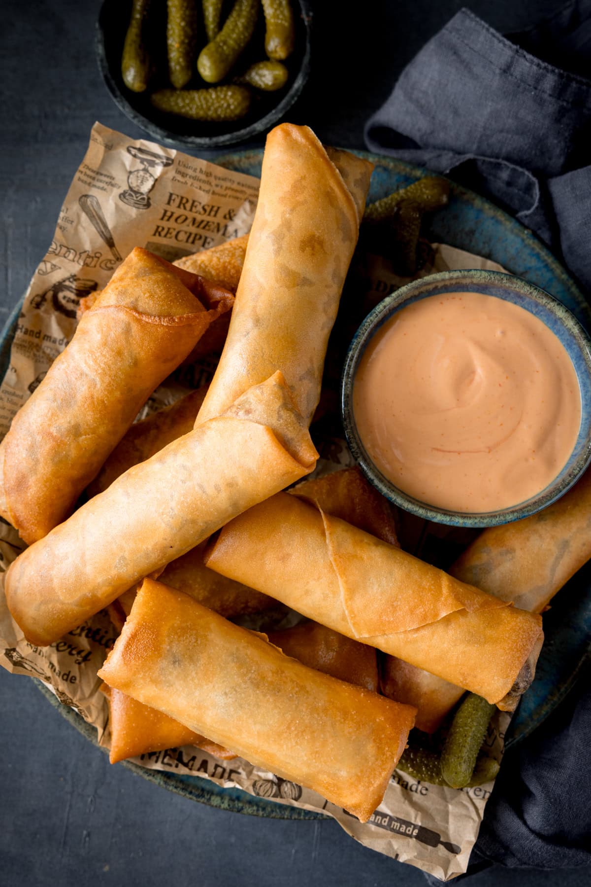 A tall, overhead shot of cheeseburger spring rolls. There are 10 cheeseburger spring rolls piled on a dark blue plate, which is lined with a light brown newspaper. There are a few pickled gherkins nestled around the spring rolls. On the right of the dish, there is a dark blue bowl filled with burger sauce. In the top left of the background, there is another dark blue bowl, filled with pickled gherkins. On the right of the background, you can see a navy blue napkin tucked underneath the main dish. This is all set on a blue background.