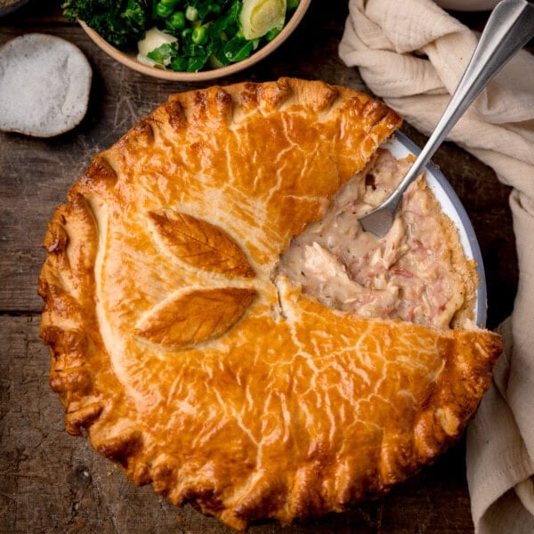 A square, overhead shot of the chicken and ham pie, with a slice taken out of it and a silver serving utensil in the filling where the piece was. In the top left corner of the image, there is a small white dish filled with sea salt flakes. To the right of that, there is a light brown bowl filled with steamed greens. In the right side of the image, there is a cream-coloured napkin tucked around the main pie dish. This is all set on a dark wooden background.