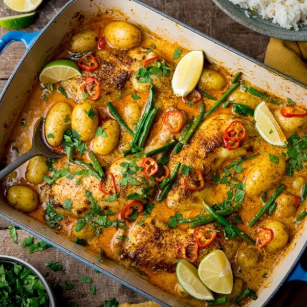 A square, overhead image of Chicken and Potato Curry. The Curry is in a large blue pan with a white inside lining. The pan is set at a diagonal in the centre of the image. There is a silver spoon sticking out of the bottom left corner of the dish. Below that, there is a grey dish filled with chopped coriander. In the top left corner of the image, you can see two lime wedges on the surface. On the right of the image, there is a yellow napkin tucked underneath the main dish and in the top right corner, there is a grey dish filled with coconut rice. There are also small bits of chopped coriander scattered around the background. This is all set on a dark, wooden background.