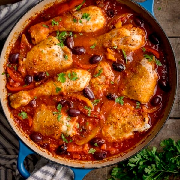 Overhead square image of chicken cacciatore in a large, shallow blue casserole dish. The dish is on a wooden table, next to some fresh herbs.