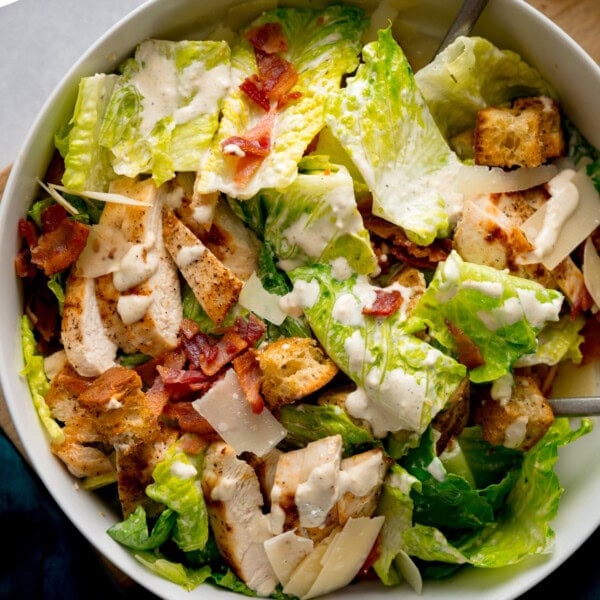 A square, overhead shot of chicken Caesar salad. The chicken Caesar salad is in a white bowl, in the top right and bottom left corners of the image, you can slightly see that the bowl is placed on a wooden board. In the bottom of the image, there is a navy blue napkin tucked beneath the bowl. This is all set on a white surface.