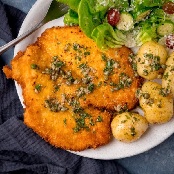 A square, overhead shot of Chicken Schnitzel. The chicken is topped with the garlic caper butter, next to it is a serving of new potatoes, and on the same plate there is a serving of butter lettuce salad and a silver fork, this is all on a large white plate. On the left side of the image, there is a navy blue napkin tucked beneath the plate. This is all set on a dark blue surface.