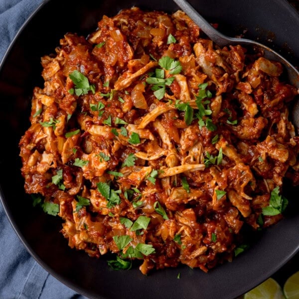 A square, overhead shot of chicken tinga in a large black bowl. The chicken is topped with sliced coriander and in the top left of the bowl, you can see a silver spoon sticking out of the dish. On the left of the background, there is a light blue napkin tucked underneath the dish, and in the bottom right corner of the image, you can slightly see two lime wedges on the surface.