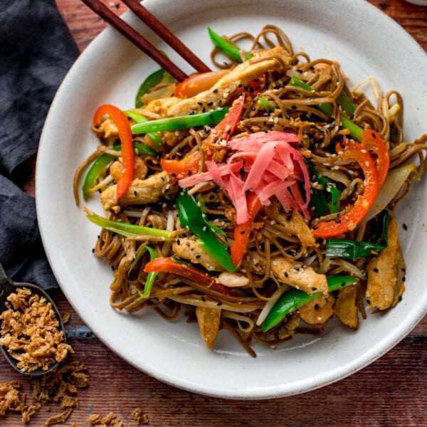 a square image of chicken yakisoba on a white plate with some chopsticks on the side, placed on a wooden board with some ingredients in the background